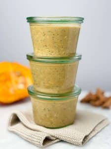 Three glass jars stacked and filled with pumpkin overnight oats with chia, set on a beige cloth with pumpkin and cinnamon sticks blurred in the background.