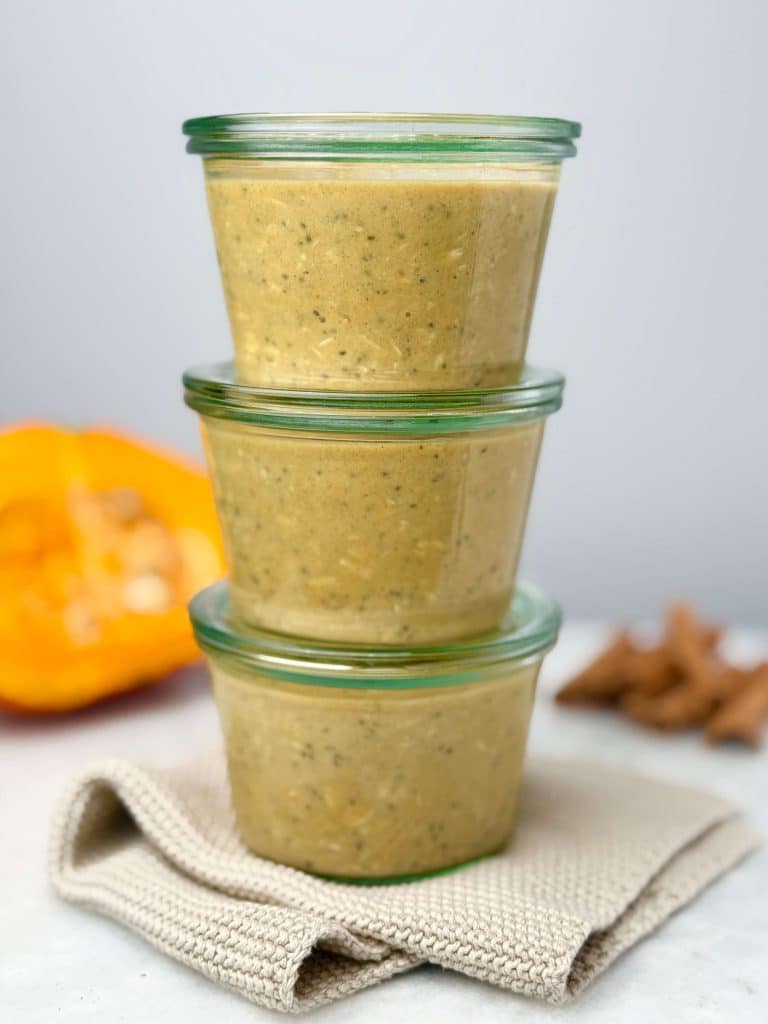Three glass jars stacked and filled with pumpkin overnight oats with chia, set on a beige cloth with pumpkin and cinnamon sticks blurred in the background.