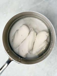 Raw chicken breasts simmering in a saucepan of water while poaching on the stovetop.