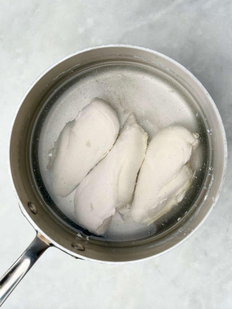 Raw chicken breasts simmering in a saucepan of water while poaching on the stovetop.