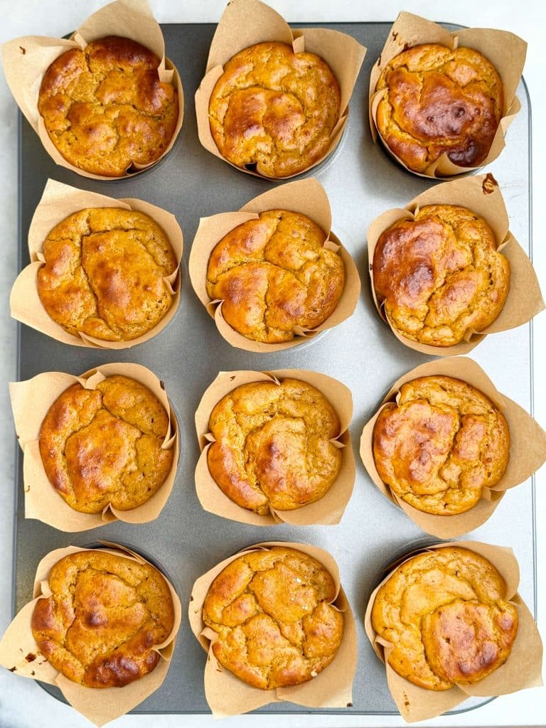 Freshly baked high protein pumpkin muffins in brown tulip liners cooling in the muffin tin with golden, slightly cracked tops.