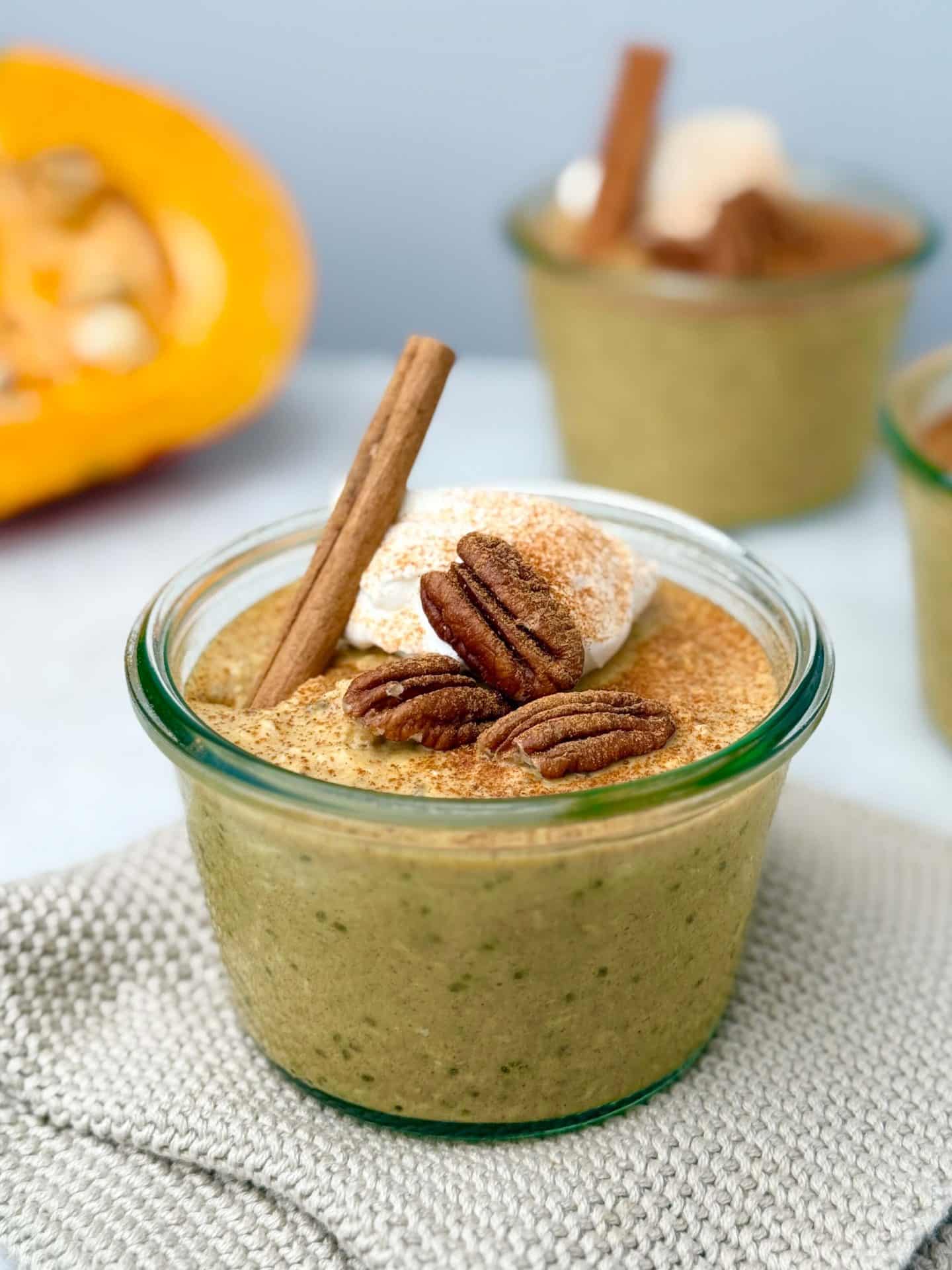 A glass jar filled with pumpkin overnight oats topped with greek yogurt, pecans, and a cinnamon stick, set on a beige cloth with a pumpkin in the background.