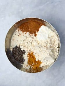 A mixing bowl filled with ingredients for pumpkin overnight oats, including chia seeds, pumpkin puree, protein powder, Greek yogurt, cinnamon, and milk, before being stirred together.