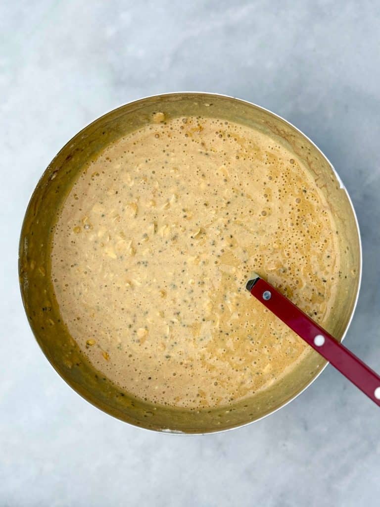 A metal bowl with a red spoon showing the pumpkin overnight oats mixture after being stirred, smooth and creamy with visible chia seeds.
