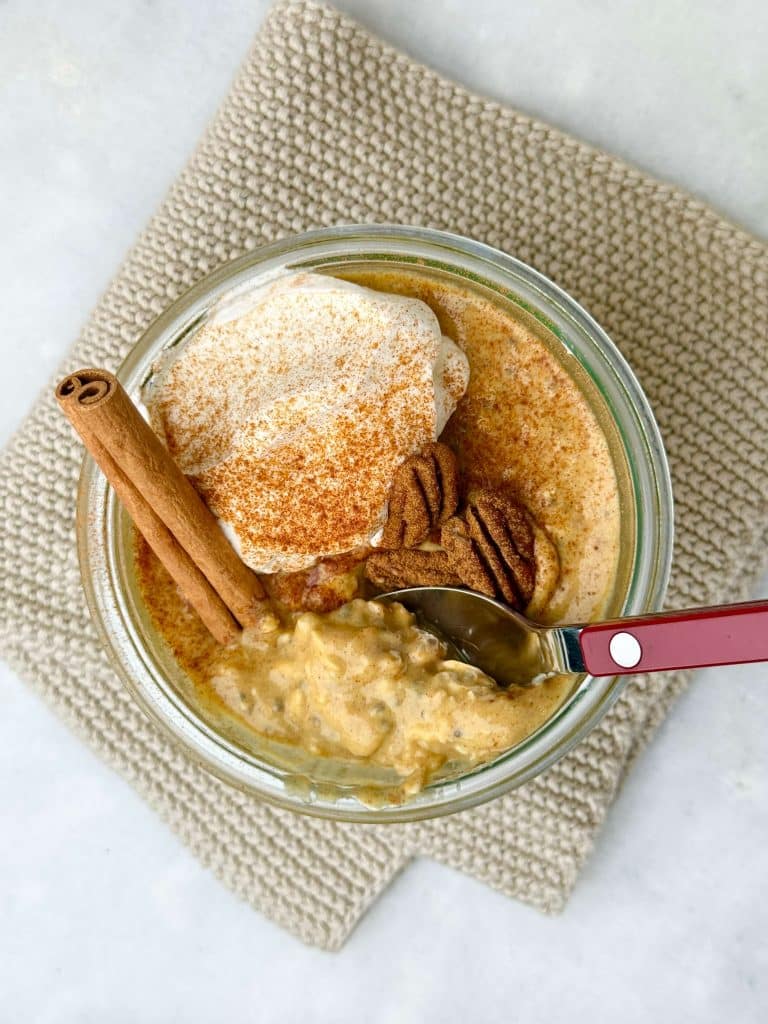 Overhead shot of pumpkin overnight oats in a glass jar, topped with greek yogurt, pecans, and a cinnamon stick, with a spoon dipped in.