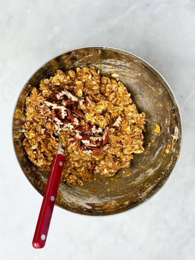 Mixing bowl of pumpkin protein ball dough with chopped pecans on top and a red-handled spoon.