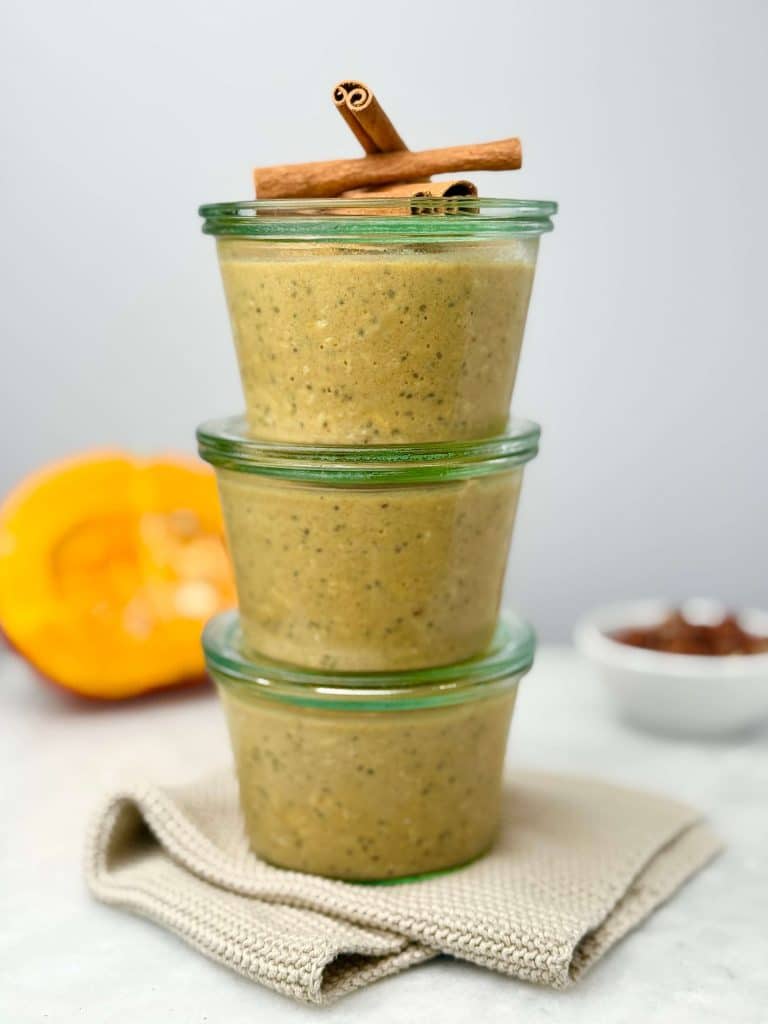 Three glass jars stacked and filled with pumpkin overnight oats, garnished with cinnamon sticks, with pumpkin and pecans blurred in the background.