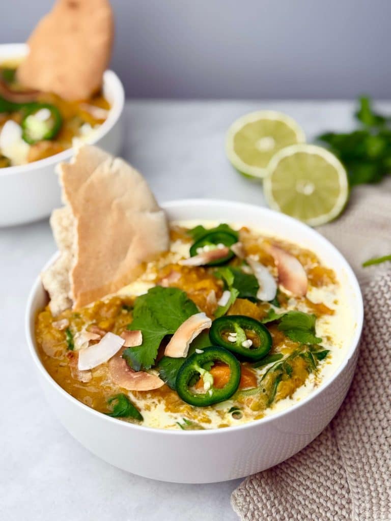 Side view of crockpot butternut squash curry served in a white bowl with cilantro, jalapeños, toasted coconut, and pita bread.