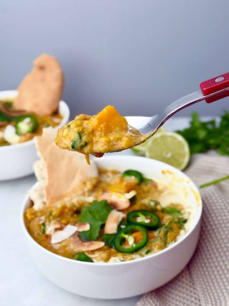 Spoon lifting a bite of creamy butternut squash curry with lentils, spinach, and coconut milk from a bowl.
