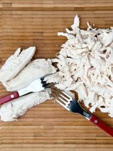 Poached chicken shredded on a wooden cutting board with two forks.