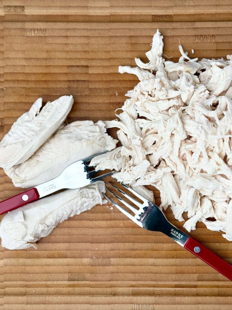 Poached chicken shredded on a wooden cutting board with two forks.