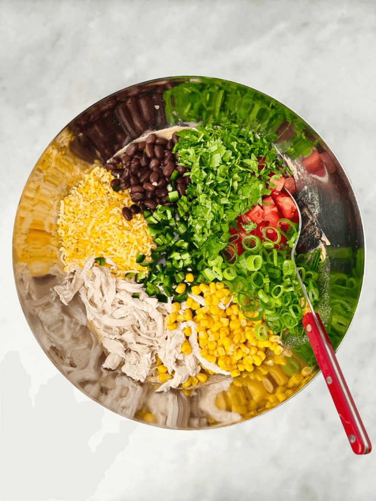 Overhead view of Southwest Chicken Salad ingredients in a mixing bowl including chicken, beans, corn, cheese, cilantro, tomatoes, jalapeños, and green onions.