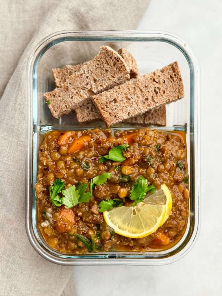 Meal prep container filled with homemade lentil soup topped with parsley and lemon, served with slices of whole grain bread.