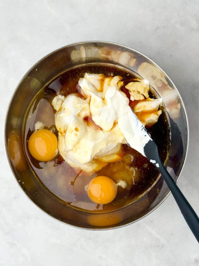 Wet ingredients including Greek yogurt, applesauce, eggs, and maple syrup in a mixing bowl for high-protein apple muffins.