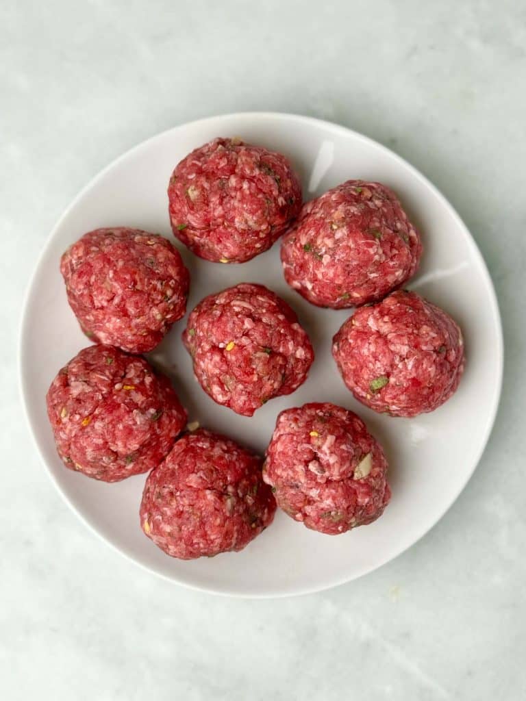 Uncooked beef sausage mixture shaped into balls on a white plate before flattening into patties.