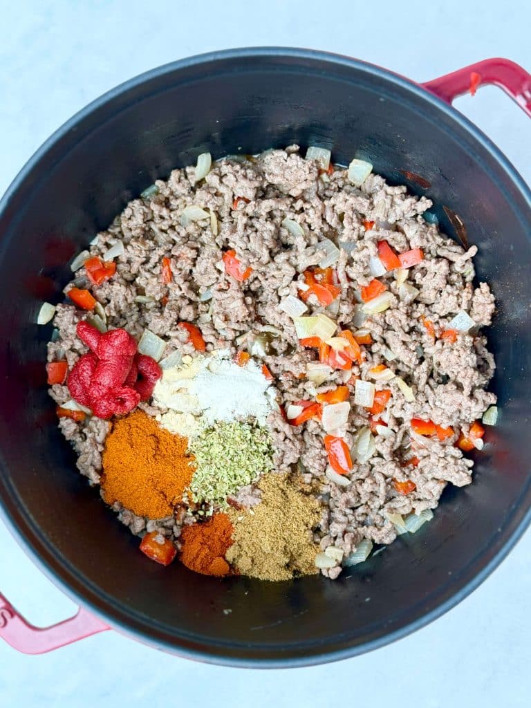 Lean ground beef cooking with onion, bell pepper, tomato paste, and spices like cumin, chili powder, and oregano in a Dutch oven.
