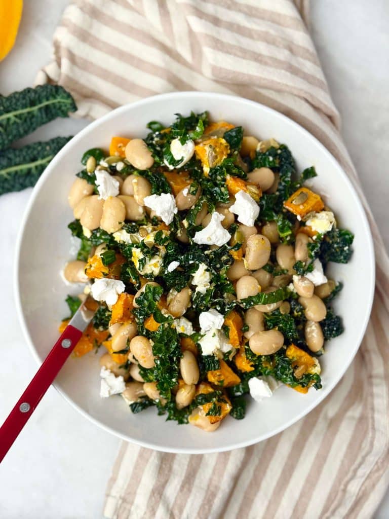 Warm butter bean salad with kale, roasted pumpkin, and goat cheese served in a white bowl with a red-handled fork.