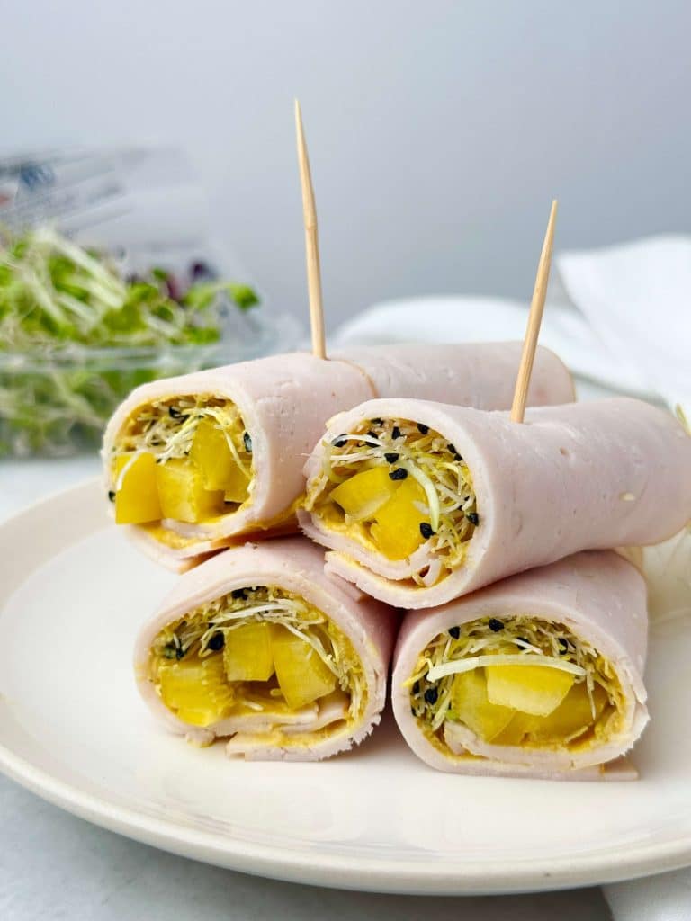 Turkey roll-ups filled with yellow bell pepper strips, fresh sprouts, and a smear of mustard, secured with toothpicks and stacked on a white plate.