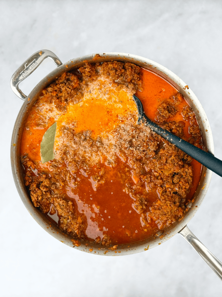 Bolognese sauce simmering in a pot with milk, chicken stock, and bay leaf for depth of flavor.