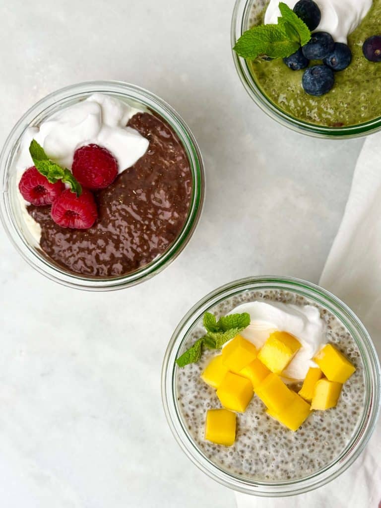 Three flavors of dairy free chia seed pudding in glass jars topped with whipped coconut cream, fruit, and mint — chocolate with raspberries, matcha with blueberries, and coconut mango with diced mango.