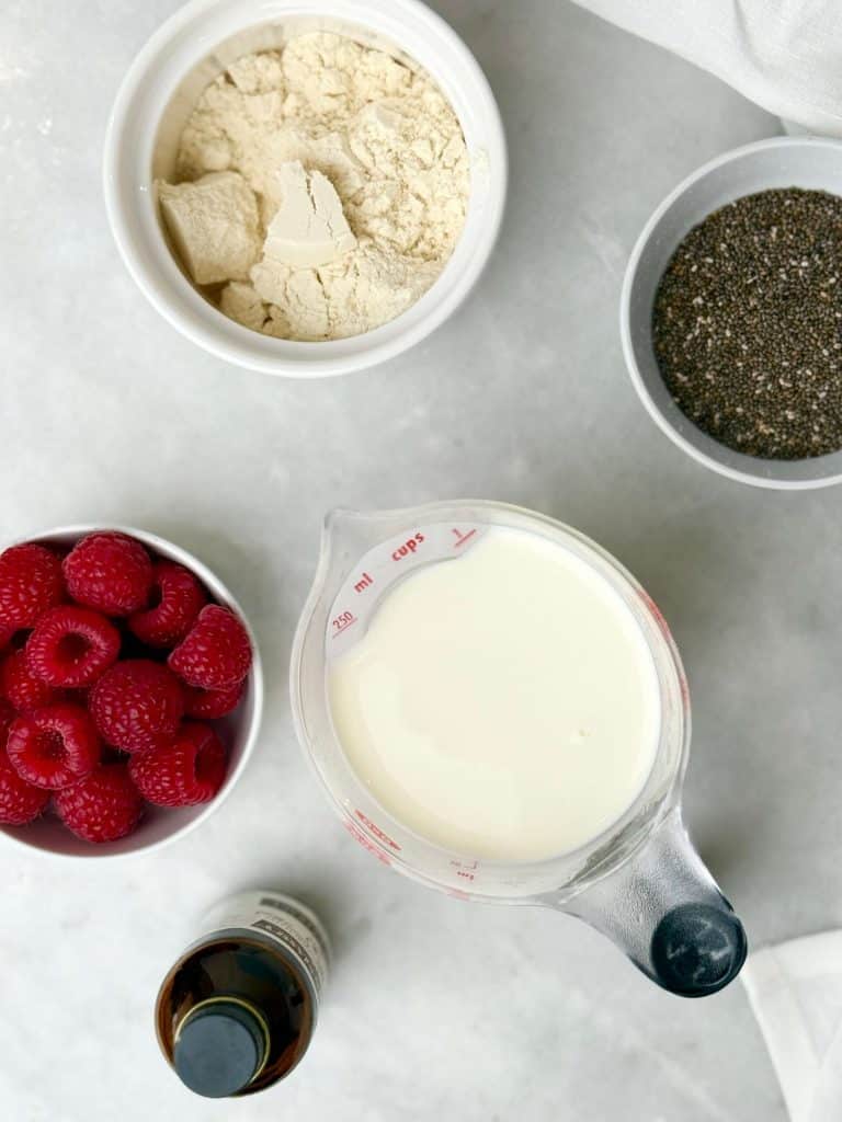Ingredients for high protein dairy free chia seed pudding including chia seeds, plant-based protein powder, almond milk, vanilla extract, and fresh raspberries.