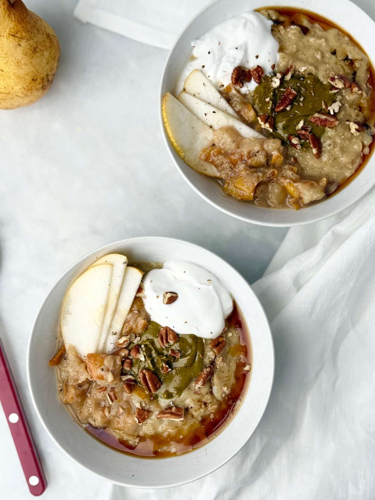 Two bowls of dairy-free protein oatmeal on white linen, garnished with pears, yogurt, nut butter, and pecans for a high-protein breakfast.