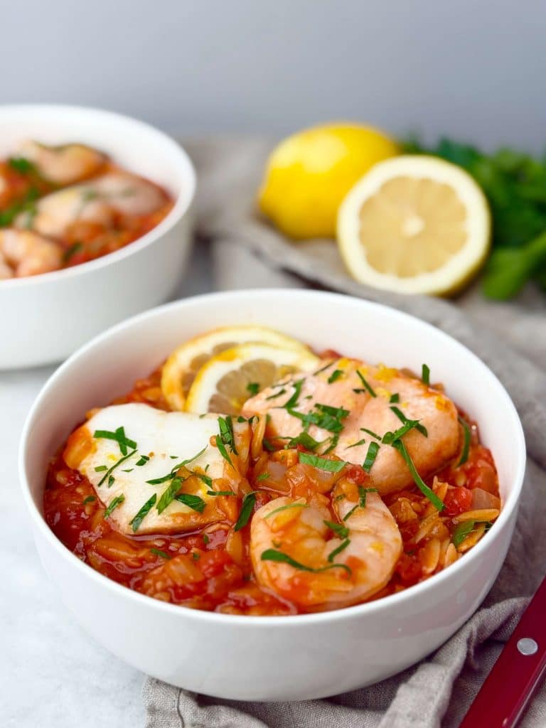 Mediterranean poached fish in tomato sauce served in a white bowl with shrimp, lemon, and parsley, photographed with fresh lemons in the background.
