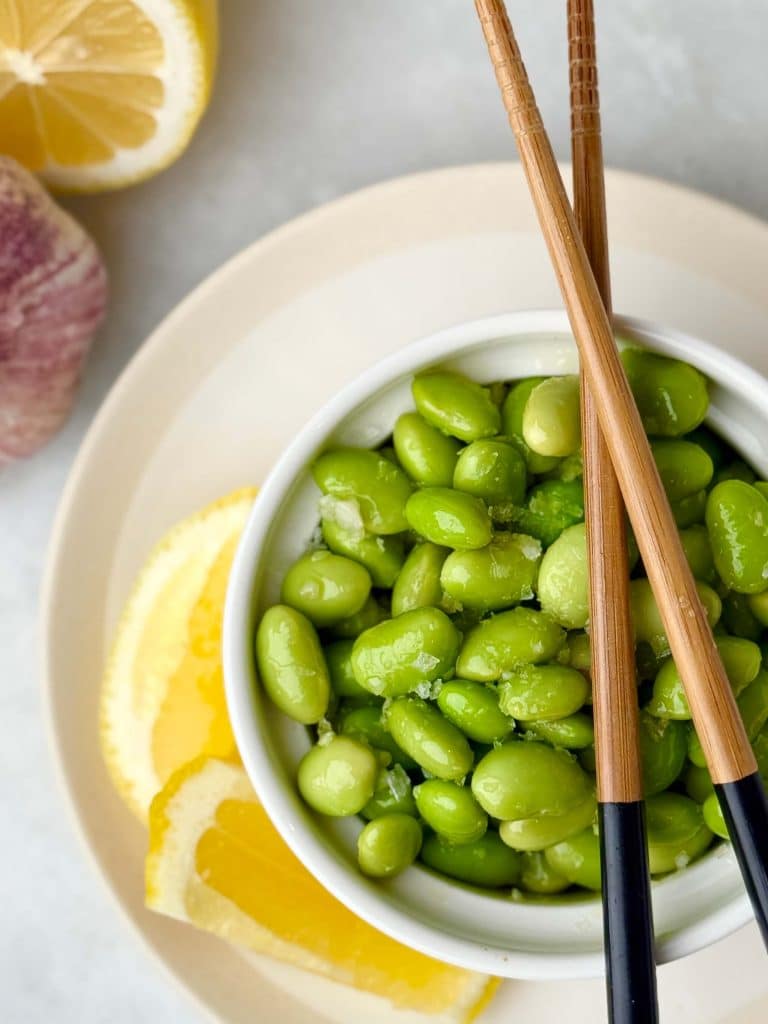 Small white bowl filled with bright green edamame sprinkled with sea salt, served with lemon wedges and chopsticks on a neutral plate.