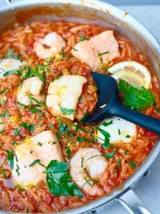 Mediterranean poached fish with shrimp and couscous in a spicy tomato sauce, garnished with parsley and lemon zest in a stainless steel pan.