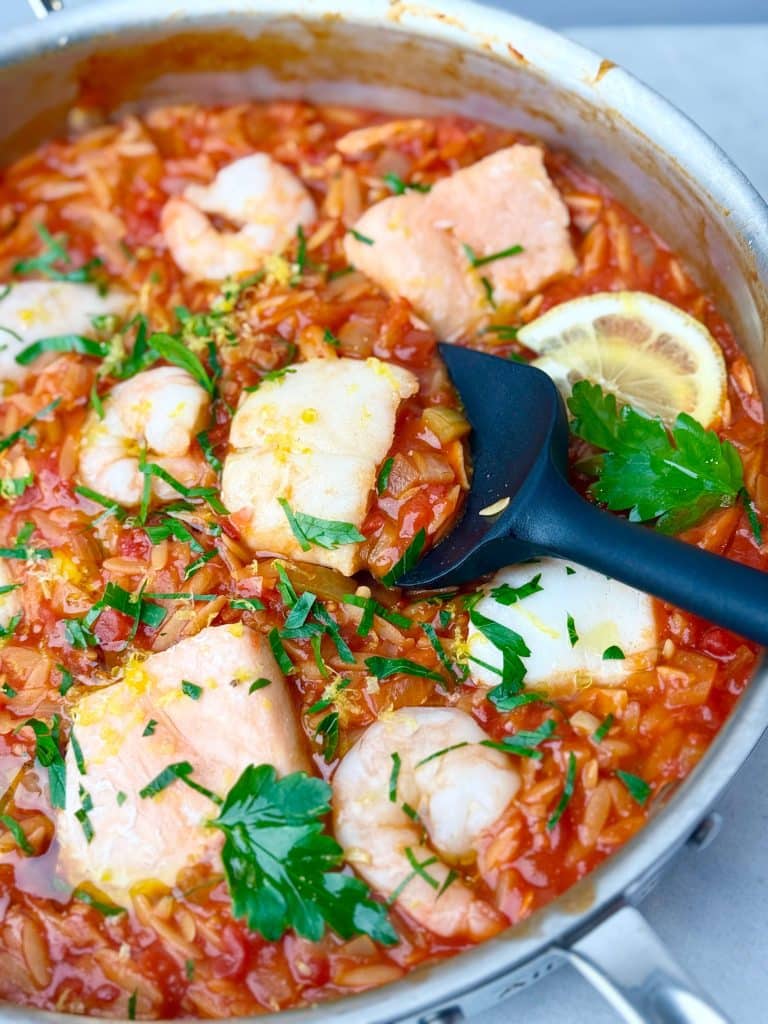Mediterranean poached fish with shrimp and couscous in a spicy tomato sauce, garnished with parsley and lemon zest in a stainless steel pan.