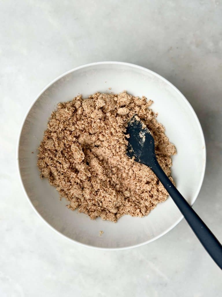 Protein cinnamon crumble mixture in a white bowl with a spatula for topping apple muffins.