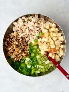 Ingredients for apple walnut chicken salad in a bowl including diced chicken, apples, celery, green onions, and toasted walnuts.