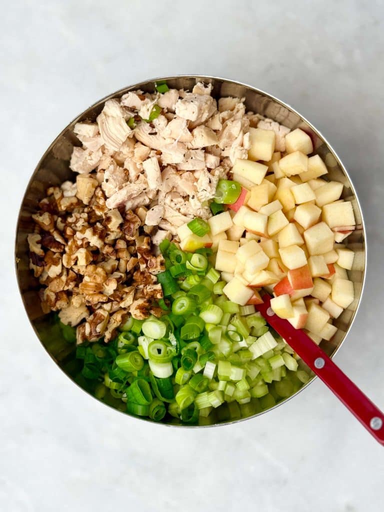 Ingredients for apple walnut chicken salad in a bowl including diced chicken, apples, celery, green onions, and toasted walnuts.