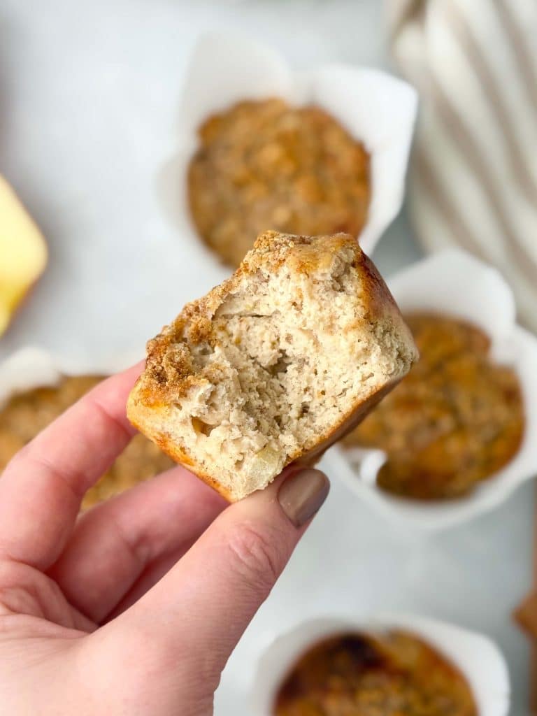 Hand holding a healthy apple cinnamon muffin with a bite taken out, showing tender crumb and apple chunks.