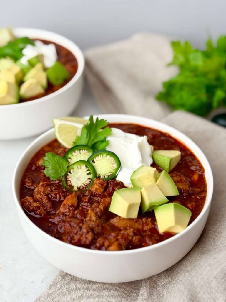 Healthy chicken chili topped with avocado, sour cream, jalapeño slices, cilantro, and lime in a white bowl.