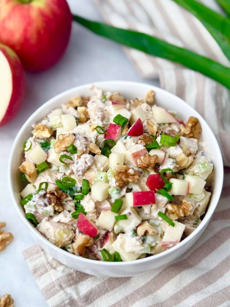 Apple walnut chicken salad in a white bowl with chunks of apple, walnuts, and green onions on a striped napkin.