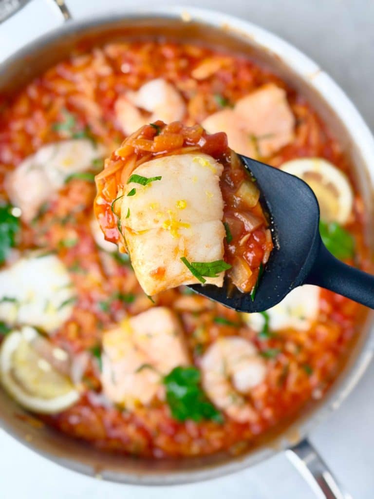 Close-up of a spoon lifting a piece of white fish from Mediterranean poached fish in spicy tomato sauce with orzo.
