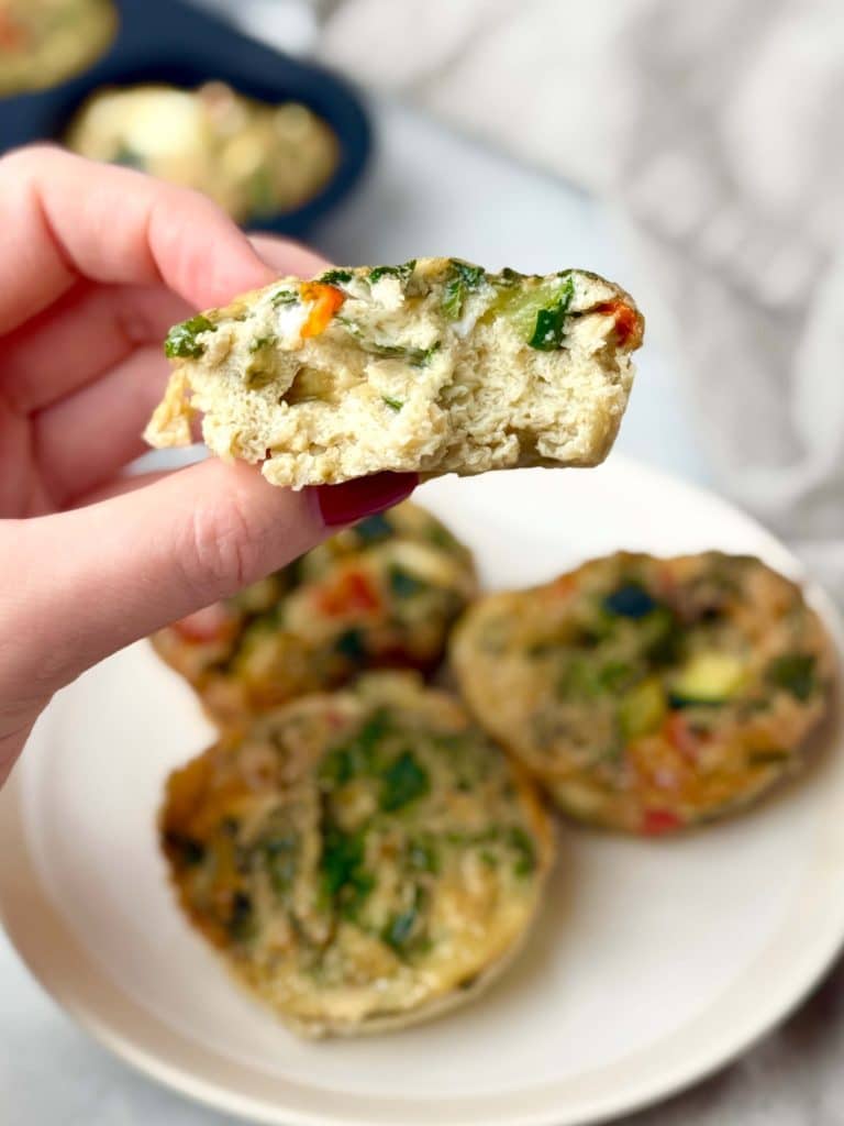 Hand holding a veggie-loaded breakfast frittata cup with visible kale, zucchini, and red bell pepper inside