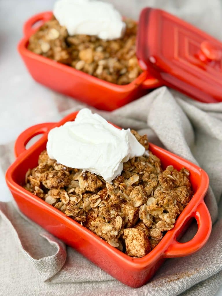 Individual red ramekins of high-protein apple crisp topped with dairy-free whipped cream.