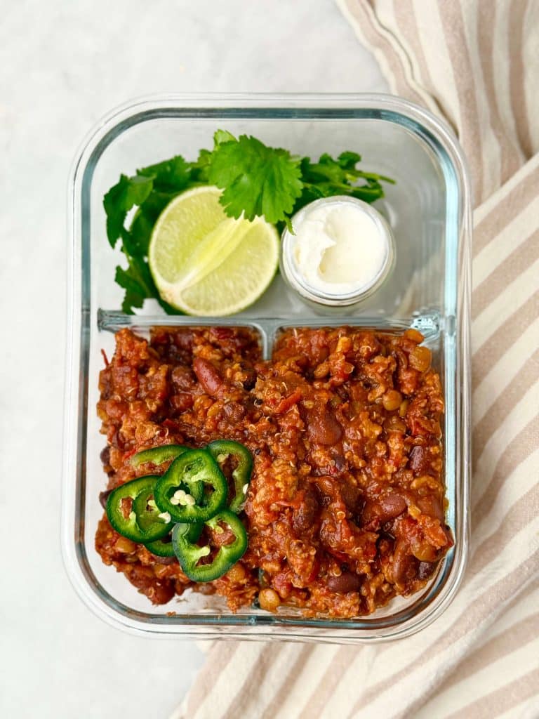 Meal prep container filled with high protein low calorie turkey chili topped with jalapeño slices, served with lime wedges, Greek yogurt, and cilantro on the side.
