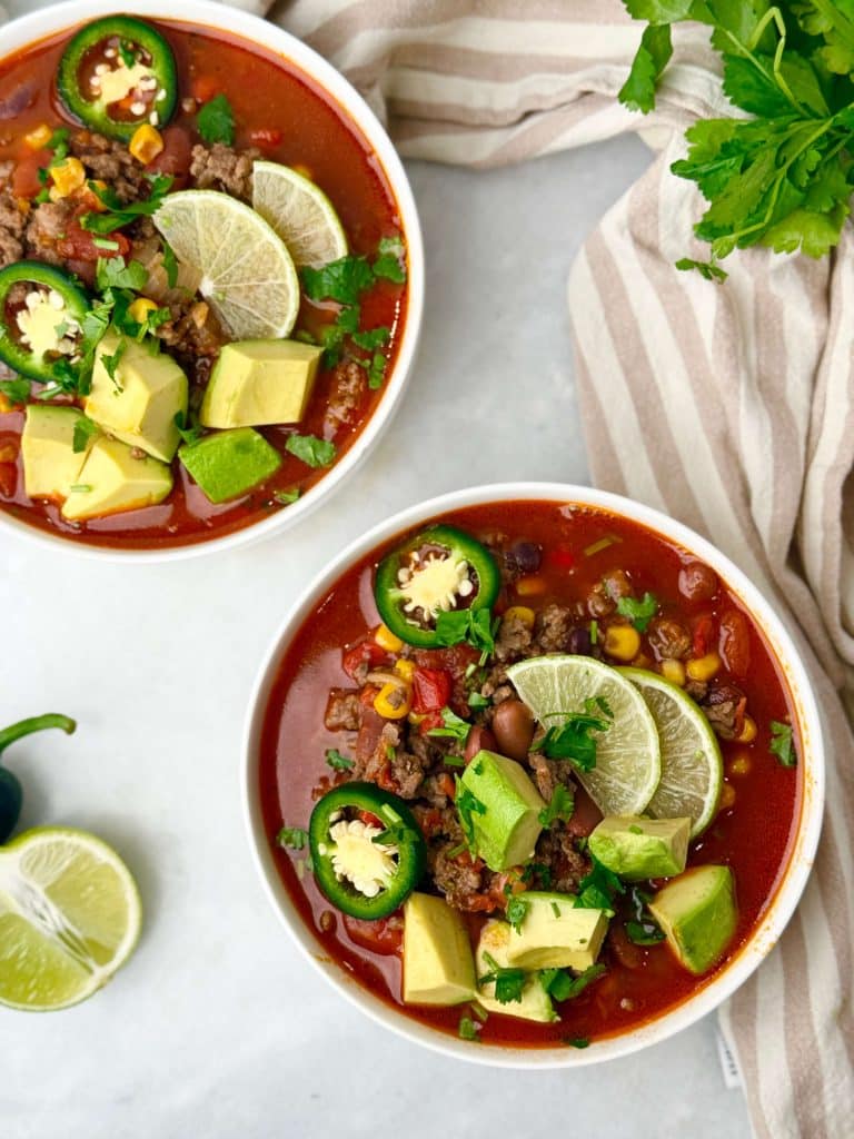 Overhead view of two bowls of high-protein beef taco soup with avocado, lime, corn, and jalapeño surrounded by fresh herbs.