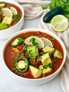 Bowl of high-protein beef taco soup topped with avocado, lime slices, jalapeño, and cilantro on a white surface.