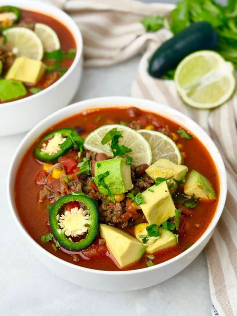 Bowl of high-protein beef taco soup topped with avocado, lime slices, jalapeño, and cilantro on a white surface.