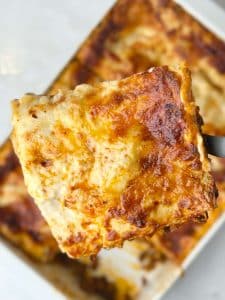 Close-up of a golden, cheesy slice of homemade lasagna lifted from the pan, showing bubbling béchamel and melted cheese.
