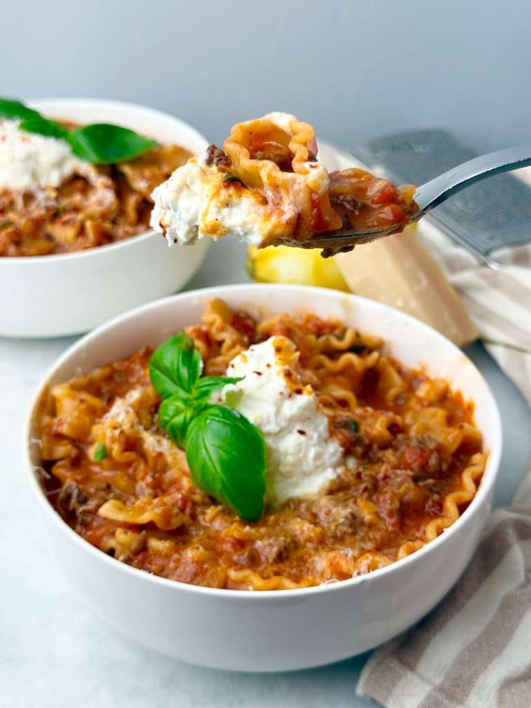 Forkful of creamy high-protein lasagna soup topped with lemon ricotta and basil, showing tender noodles, ground beef, and rich tomato sauce in a white bowl.