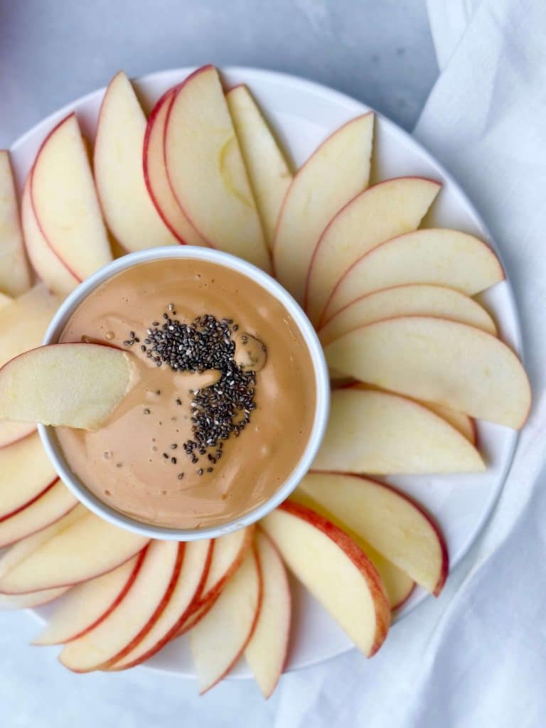 Plate of thinly sliced red apples arranged around a small white bowl of creamy peanut butter protein dip topped with chia seeds.