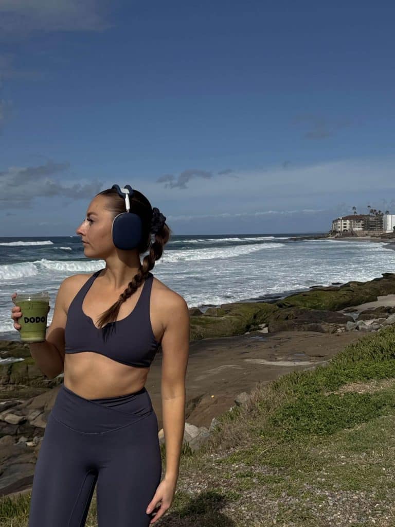 Woman drinking a green smoothie by the ocean, wearing headphones and workout clothes, embodying health and vitality.