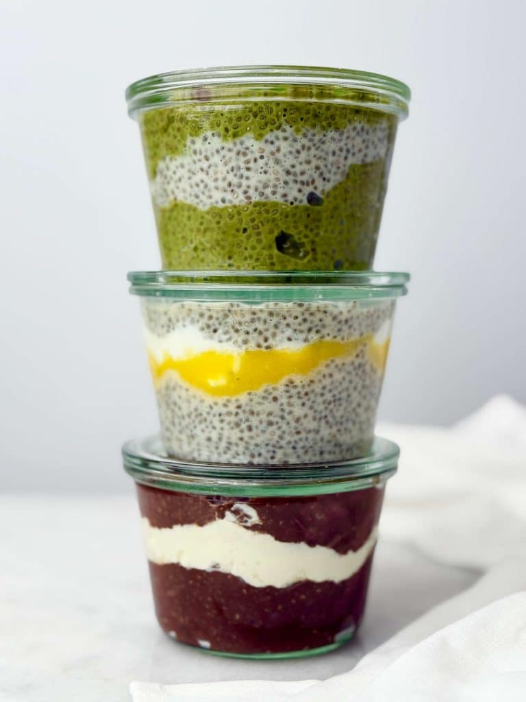 Stack of three glass jars showing different flavors of dairy free chia seed pudding — matcha with blueberries, coconut mango with purée, and chocolate with coconut cream.