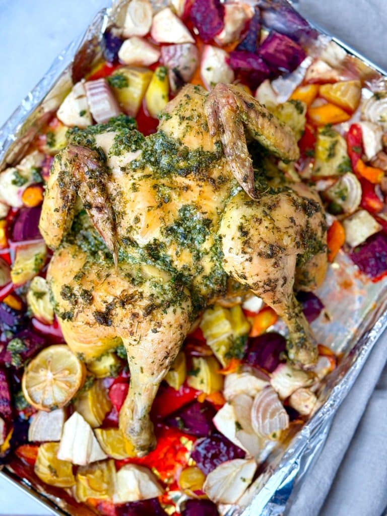 Whole oven roasted spatchcock chicken resting on a sheet pan surrounded by caramelized beets, carrots, and parsnips.