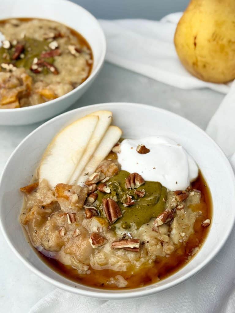 Creamy dairy-free protein oatmeal topped with pistachio butter, coconut yogurt, sliced pear, chopped pecans, and maple syrup in a white bowl on marble surface.